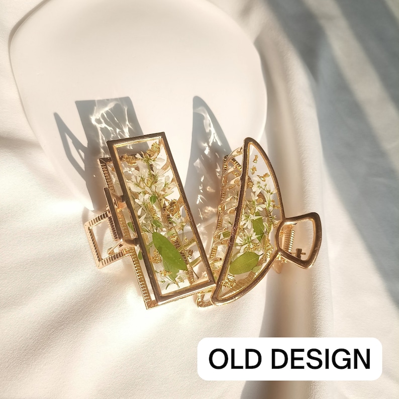 Pu&ograve; includere: Due fermagli per capelli dorati con un design floreale. I fermagli sono in metallo e hanno un inserto in resina trasparente con fiori bianchi e foglie verdi. Il testo "OLD DESIGN" &egrave; stampato sullo sfondo bianco.