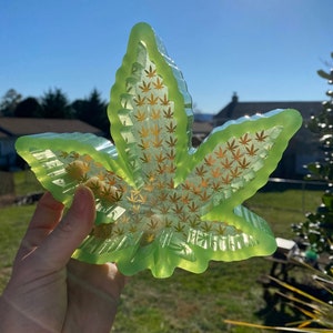 Puede incluir: Una bandeja de resina translúcida de color verde lima con forma de hoja de cannabis. La bandeja presenta un patrón de diseños de hojas de cannabis doradas. La bandeja se sostiene contra un fondo soleado al aire libre.
