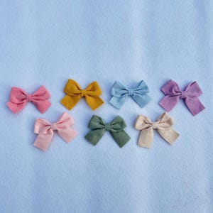 Può includere: Sette fiocchi per capelli in tessuto di vari colori, tra cui rosa, azzurro, verde, giallo e crema. I fiocchi sono disposti in fila su una superficie blu chiaro.