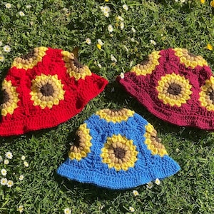 May include: Three crocheted bucket hats with sunflower designs. The hats are red, burgundy, and blue.