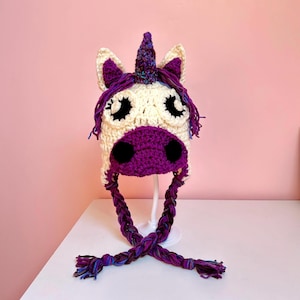 Op de afbeelding: Een gebreide unicorn-muts met een wit gezicht, paarse neus en zwarte ogen. De muts heeft lange paarse en blauwe gevlochten banden.