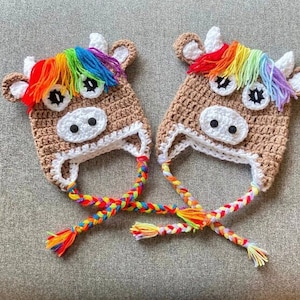 May include: Two brown crocheted cow hats with rainbow yarn accents. The hats have white felt eyes and noses. The hats have long rainbow yarn ties.
