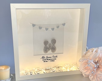 May include: A white shadow box with a light inside, featuring a pebble art family of three with a string of blue hearts above them. The box also includes the text "Alfie James Smith 10th March 2023 7lb 4oz 9:20am".