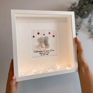 May include: A white framed picture with a white background. The picture features a pebble art design of two figures holding each other. There are red hearts hanging above the figures. The text "Celebrating 40 years of love Mum and Dad 12/03/1983" is written below the figures. The frame has a light inside and there are clear crystals at the bottom of the frame.