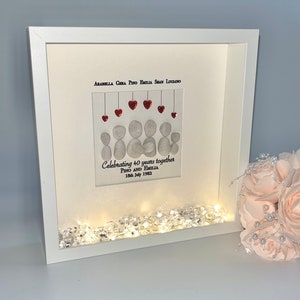 May include: A white picture frame with a white mount and a glass front. Inside the frame is a pebble art family portrait with red hearts hanging above the figures. The text "Celebrating 40 years together Pino and Emilia 18th July 1982" is written below the figures. The frame is lit with white fairy lights.