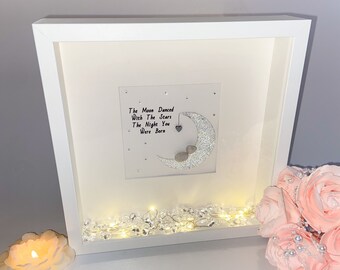May include: A white framed picture with a silver glitter moon and two small gray stones. The text in the picture reads "The moon danced with the stars the night you were born". The picture is surrounded by white fairy lights and clear crystals.