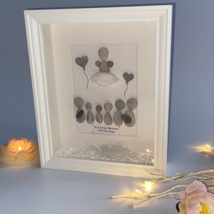 May include: A framed pebble art piece with a white background. The art depicts an angel with a halo, holding a heart-shaped balloon. Below the angel are five figures, also made of pebbles, with the text "In Loving Memory Of Our Papa" written below them. The frame is white and has a white mount.