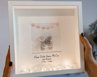 May include: A white picture frame with a light inside, displaying a pebble art family of three. The frame has a string of pink hearts across the top and a personalized inscription that reads "Eloise Violet James McCue 13th March 6.13oz 6.55cm".