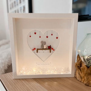 May include: A white frame with a heart-shaped opening containing a pebble art couple sitting on a bench with a red rose. The couple is made of grey and white pebbles. The heart is surrounded by red hearts hanging from strings. The frame has a white background and a white mount. The bottom of the frame has a string of white lights. The text "Happy Valentine's Day 2021" is written in black at the bottom of the frame.
