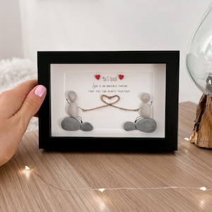 May include: A framed pebble art piece with two figures holding a string that forms a heart. The text reads "Love is an invisible thread that ties two hearts together." The names "Max & Hannah" are written above the text.