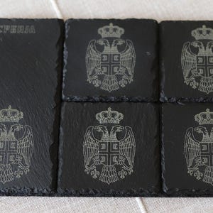 Puede incluir: Conjunto de seis posavasos de pizarra negra con un diseño grabado en blanco del escudo de armas de Serbia. Los posavasos son cuadrados y tienen una superficie rugosa y texturizada. La palabra "SRBIJA" está grabada en uno de los posavasos.