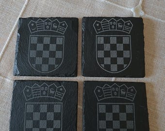 May include: A set of four square black slate coasters, each featuring an engraved Croatian coat of arms. The design includes a shield with a red and white checkerboard pattern topped by a crown containing five smaller historical shields.