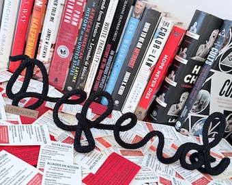 May include: A black crocheted "Defend" word art piece is displayed in front of a collection of books. The books have titles like "How to Be an Antiracist" and "White Fragility". Red and white cards with Spanish text are scattered around.