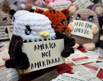 May include: A collection of handmade crochet animals, including an eagle holding a sign that reads "AMERICA NOT AMERIKKKA". Other animals are visible in the background. The sign also has the text "LATINXLOOPS".
