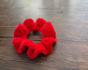 May include: A vibrant red scrunchie made of a soft, plush material. The scrunchie is circular with a gathered design, and the text "LATINXLOOPS" is visible on the surface. This hair accessory adds a pop of color.