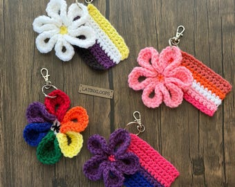 May include: Four handmade crochet keychains featuring flower designs and colorful flag patterns. The keychains include a white flower with a yellow and purple accent, a pink flower with orange and pink accents, a rainbow flower, and a purple flower with pink and blue accents.