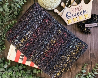 May include: A dark gray crocheted book sleeve with colorful flecks, resting on a book. A wooden crown with the text "YARN Queen" and a ball of yarn are also visible. The sleeve is handmade and designed to protect books.