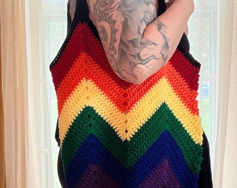 May include: A handmade crochet tote bag with a vibrant rainbow chevron pattern. The bag features a black border and straps, and the colorful design includes red, orange, yellow, green, blue, and purple. The bag is a stylish accessory.