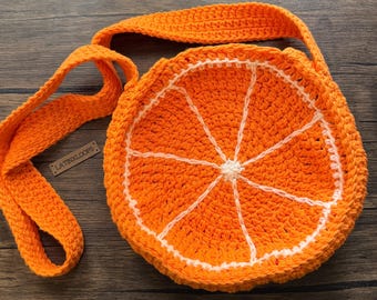 May include: A round, crocheted crossbody bag shaped like an orange slice, featuring vibrant orange yarn with white yarn detailing to represent the segments and pith. A small rectangular wooden tag labeled LATINXLOOPS is attached to the strap.