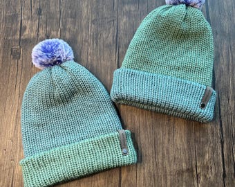 May include: Two knitted beanies in shades of teal and blue, each topped with a fluffy purple pom-pom. The hats have a folded brim and a small tag with text. The hats are displayed on a wooden surface.