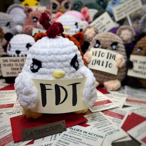 May include: A collection of handmade crochet plushies, including a white chicken holding a sign that reads "FDT." Other plush animals are visible in the background, some holding signs. The scene is set on a surface covered with printed cards.