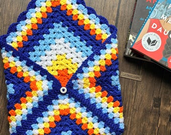 May include: A handmade crochet envelope-style case in a granny square pattern. The case features a button closure and a vibrant color scheme of blue, white, yellow, and orange. The text "LATINXLOOPS" is visible on the lower right.