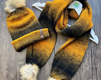 May include: A knitted hat and scarf set in a gradient of black, yellow, and brown. The hat has a faux fur pom-pom and a folded brim. The scarf has matching faux fur pom-poms at the ends. The set is displayed on a white hanger.