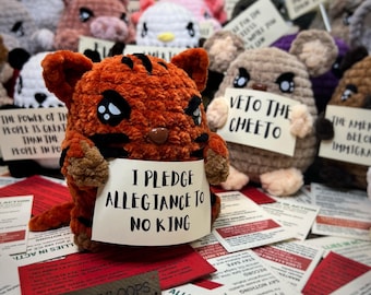 May include: A collection of handmade crochet animals, including a tiger holding a sign that reads "I Pledge Allegiance to No King." Other animals hold signs with text. The tiger is orange with black stripes. The background includes printed cards.