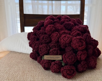 May include: A handmade bouquet of burgundy crochet roses. The flowers are densely packed together, creating a full, textured appearance. A small wooden tag with the text "LATINXLOOPS" is attached to the bouquet. The bouquet rests on a beige surface.
