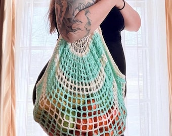 May include: A crocheted net bag in white and light teal, filled with various fruits. The bag has a rounded top and a wide, open weave design. The fruits inside are a mix of red, orange, and green, creating a colorful display. The bag is held by a person.