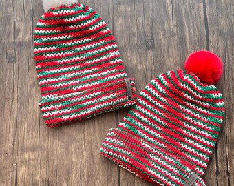 May include: Two knitted beanies with red, green, and white horizontal stripes. One beanie has a white pom-pom, and the other has a red pom-pom. The beanies have a folded cuff with a small leather tag.