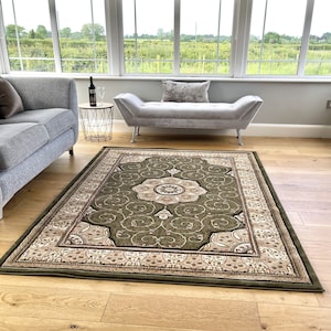May include: A green and cream patterned rug with a floral design in the centre. The rug is laid out on a wooden floor in a room with a sofa and a bench.