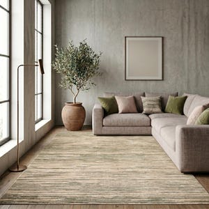 May include: A rectangular area rug featuring a horizontal striped pattern in shades of beige, tan, and muted sage green. The rug has a low-pile texture and covers a wooden floor, extending from the foreground towards a large sectional sofa.