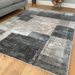 May include: A grey and beige area rug with a geometric pattern. The rug has a modern design with squares and rectangles in different shades of grey and beige.