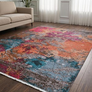 May include: A rectangular area rug featuring an abstract pattern with vibrant splashes of magenta, bright orange, deep teal, and muted grey tones. The rug has a short pile texture and a white fringed edge along the shorter side of the carpet.