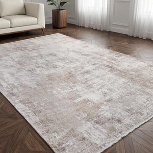 May include: A rectangular area rug featuring a distressed, abstract pattern in shades of cream, beige, and soft brown. The rug has a low-pile texture with a subtle, weathered appearance and finished edges, covering a portion of a dark wood floor.
