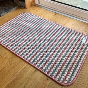 May include: A pink and grey striped doormat with a textured surface. The doormat has a pink border.