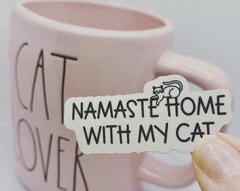 May include: White sticker with black text that reads "Namaste Home With My Cat" and a small black cat illustration. The sticker is on a pink mug with the word "Cat" written on it.