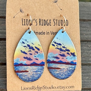 May include: A pair of teardrop-shaped earrings with a painted sunset scene over a lake. The earrings are made of metal and have a gold-tone finish. The earrings are on a card that says "Lion's Ridge Studio" and "Made in Vermont".