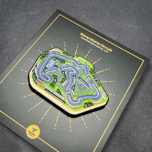 May include: A black enamel pin featuring a detailed map of the Silverstone Circuit in Northamptonshire, UK. The pin is surrounded by a gold starburst design and the text "Silverstone Circuit" and "Northamptonshire, UK".