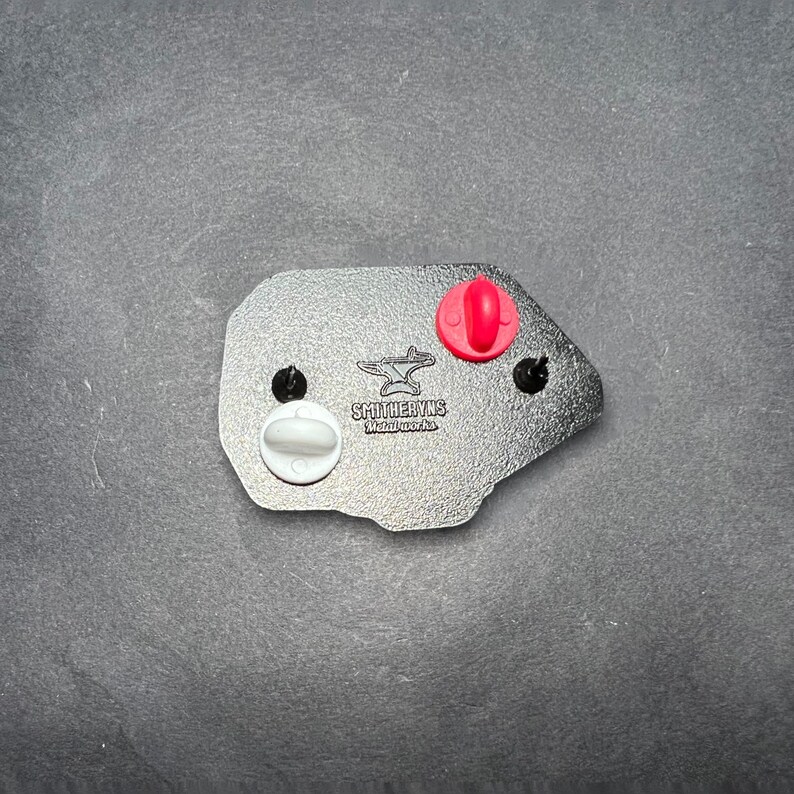 May include: Black and white enamel pin featuring a blacksmith's anvil with the text "Smitheryns Metalworks". The pin has a red rubber backing and a white rubber backing.