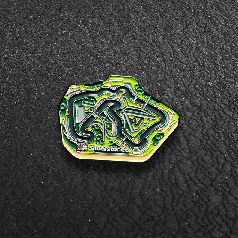 May include: A green and black enamel pin shaped like the Silverstone racetrack. The pin has a yellow border and the text "Silverstone" in white.