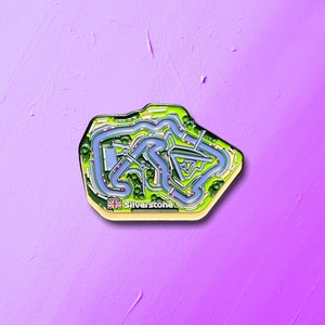 May include: A black and green enamel pin shaped like the Silverstone racetrack. The pin features a blue track with green grass and trees. The text "Silverstone" is printed on the pin.