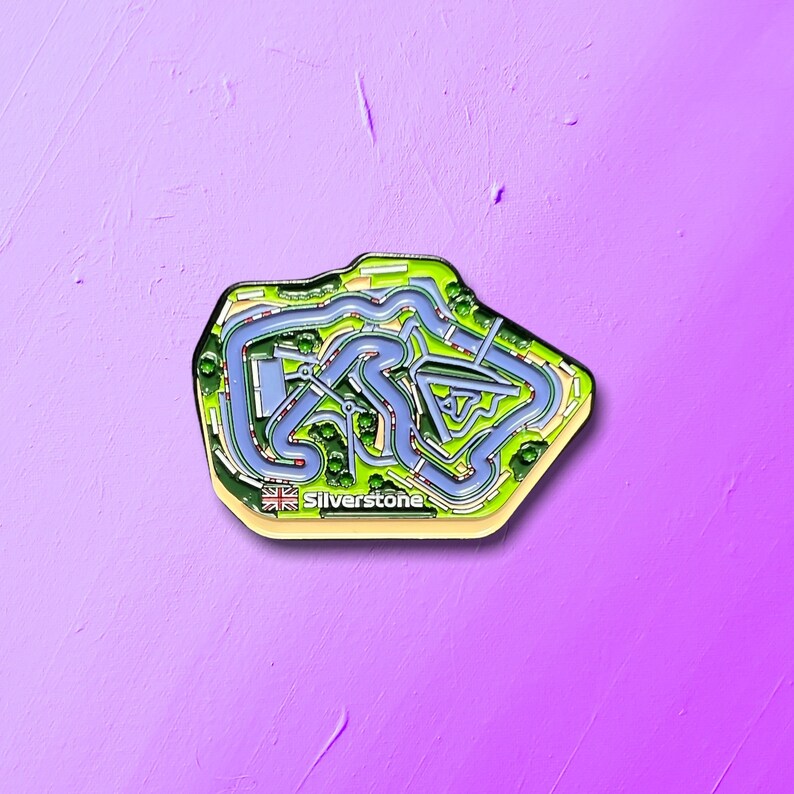 May include: A black and green enamel pin shaped like the Silverstone racetrack. The pin features a blue track with green grass and trees. The text "Silverstone" is printed on the pin.