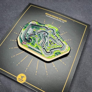 May include: A black and green enamel pin depicting the Silverstone Circuit race track in Northamptonshire, UK. The pin features the text "Silverstone Circuit" and "Silverstone" with a Union Jack flag.