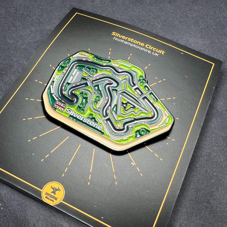 May include: A black and green enamel pin depicting the Silverstone Circuit race track in Northamptonshire, UK. The pin features the text "Silverstone Circuit" and "Silverstone" with a Union Jack flag.