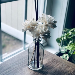 Pode incluir: Uma garrafa de vidro transparente com um arranjo de flores brancas e varetas difusoras pretas. A garrafa está sobre uma mesa de madeira.