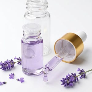 May include: A clear glass bottle and a second glass bottle filled with purple liquid, featuring a gold-toned dropper cap with a white top. A single drop of purple liquid hangs from the glass pipette, surrounded by sprigs of purple lavender flowers.