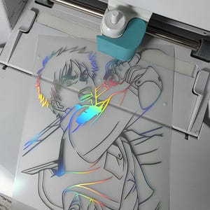 May include: A Cricut Joy cutting machine processing a sheet of holographic vinyl featuring a detailed line art illustration of an anime character with spiky hair, holding a sword, with the text Cricut Joy visible on the machine housing.