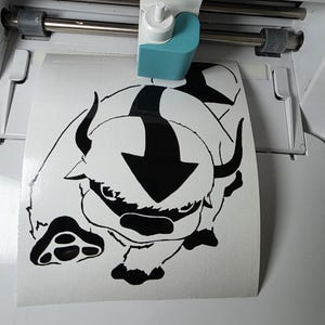 May include: A white Cricut Joy cutting machine with a teal and white blade housing. A sheet of white adhesive material is being fed through the machine, featuring a black vinyl decal of a stylized bison character with a large arrow on its head.